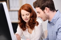 Attractive young man and woman working together sitting at a desk with a desktop computer reading paperwork together with a smile-1