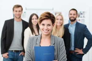 Group of job applicants with a smiling confident young woman in the foreground holding her Curriculum vitae in her hands